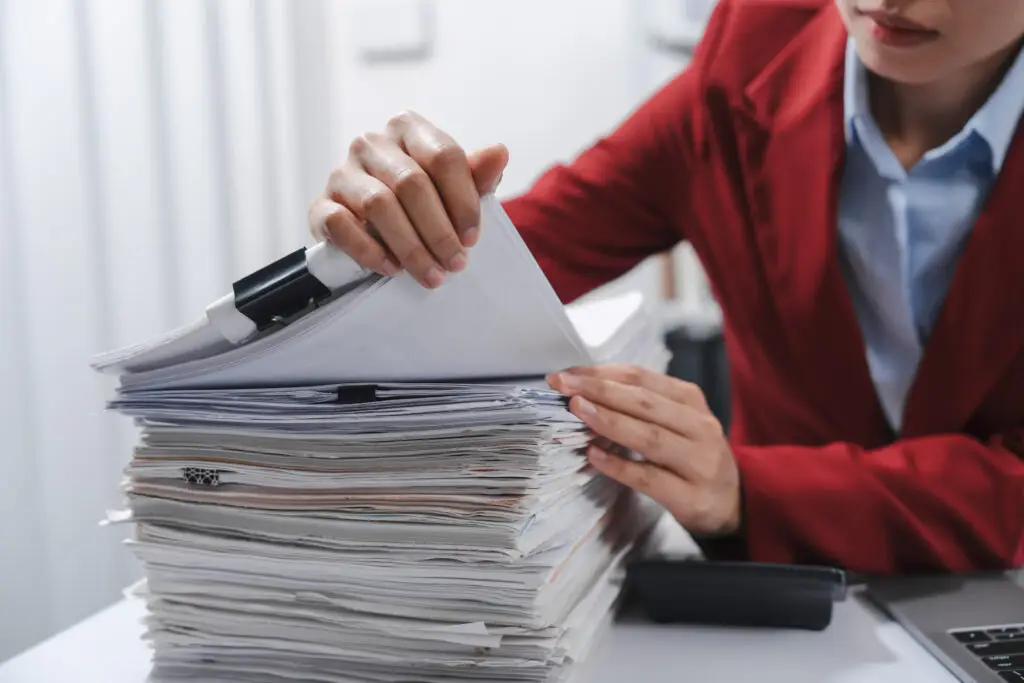 Close up hands of person in a red blazer organizing a substantial stack of various papers, likely sorting or reviewing documents for yearly tax purposes