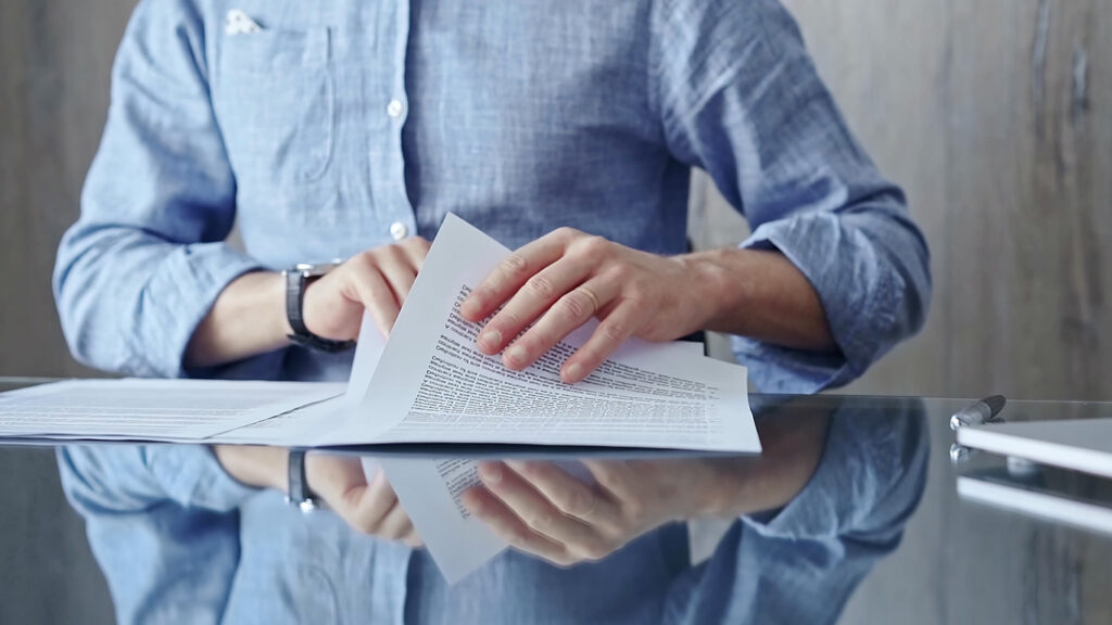 Businessperson analyzing document at desk. Close-up of a professional auditor or lawyer reviewing a lengthy paper report in office setting